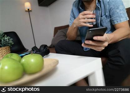 designer man hand using mobile payments online shopping,omni channel,drinking water,sitting on sofa in living room,green apples in wooden tray