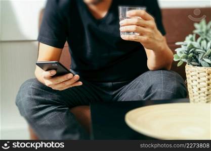 designer man hand with glass of water using smart phon for mobile payments online shopping,omni channel,sitting on sofa in living room,vase rattan with plant and wooden tray on table