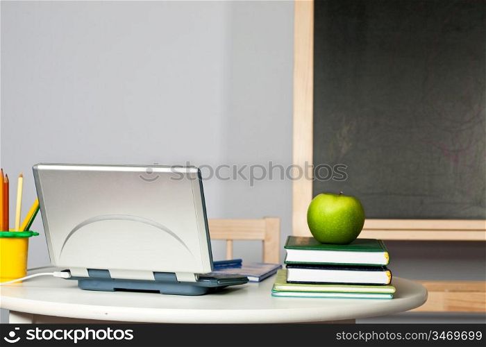 Desk with apple, computer and textbook in class against blackboard. Focus on computer