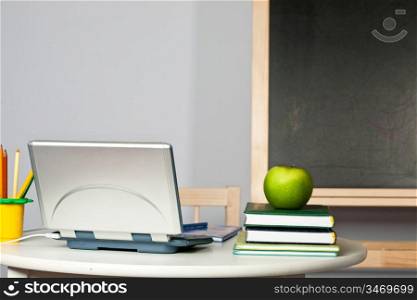 Desk with apple, computer and textbook in class against blackboard. Focus on computer