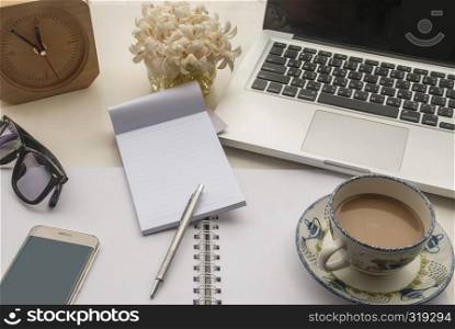 Desk with laptop, notebook, glasses, clock and flowers on a white table