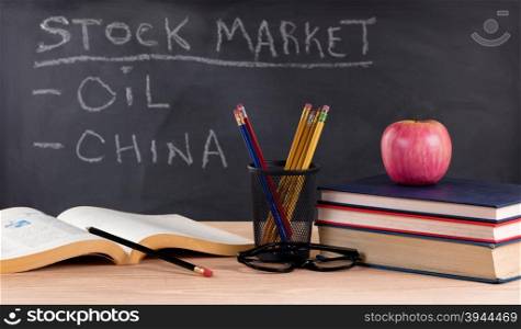 Desktop with books, pencils, apple, reading glasses and blackboard in background with stock market concept. Selective focus on front part of desk objects.