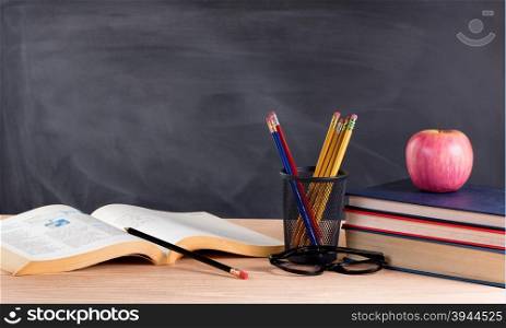 Desktop with books, pencils, apple, reading glasses and blank blackboard in background. Selective focus on front part of desk objects.