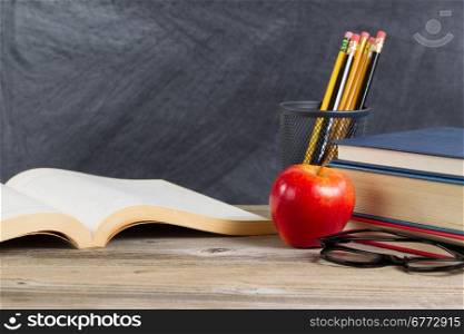 Desktop with books, red apple, reading glasses, and pencils in front of blackboard. Layout in horizontal format with plenty of copy space.