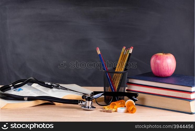 Desktop with stethoscope, medicine pills, books, pencils, apple, reading glasses and blank blackboard in background. Selective focus on front part of desk objects.