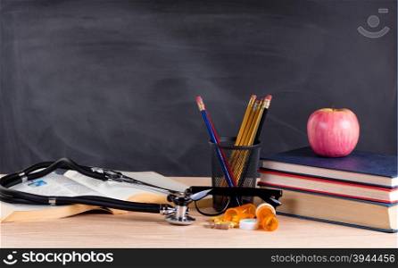 Desktop with stethoscope, medicine pills, books, pencils, apple, reading glasses and blank blackboard in background. Selective focus on front part of desk objects.