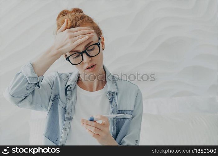 Desperate unhappy young woman sad because of infertility keeps hand on forehead looks at pregnancy test with negative result cannot have baby for long time wears eyeglasses and denim jacket.. unhappy young woman sad because of infertility looks at pregnancy test with negative result