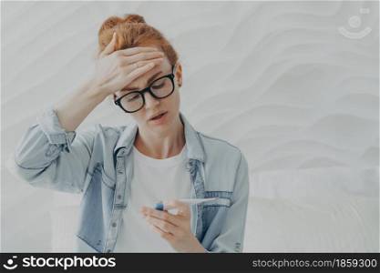 Desperate unhappy young woman sad because of infertility keeps hand on forehead looks at pregnancy test with negative result cannot have baby for long time wears eyeglasses and denim jacket.. unhappy young woman sad because of infertility looks at pregnancy test with negative result