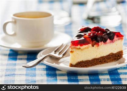 Dessert background - fruit cheese cake on plate with fork and coffee cup on blue checkered tablecloth. Cake on plate with fork and coffee cup
