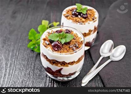 Dessert Black Forest of cherries, chocolate biscuit and soft cottage cheese with cream in two glasses, a towel, mint and a spoon on dark wooden board background