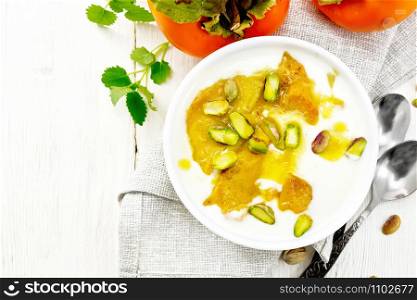 Dessert of yogurt, persimmon and honey with vanilla, cardamom and pistachios in a bowl on a napkin, mint and spoon on wooden board background from above