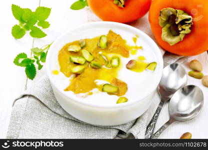 Dessert of yogurt, persimmon and honey with vanilla, cardamom and pistachios in a bowl on a napkin, mint and spoon on background of light wooden board