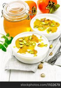 Dessert of yogurt, persimmon and honey with vanilla, cardamom and pistachios in two bowls on a napkin, mint and spoon on background of light wooden board