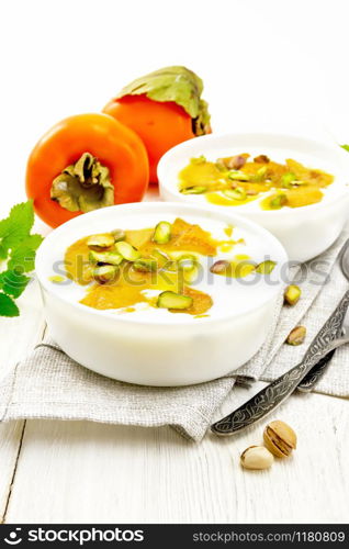 Dessert of yogurt, persimmon and honey with vanilla, cardamom and pistachios in two bowls on a towel, mint and spoon on background of light wooden board