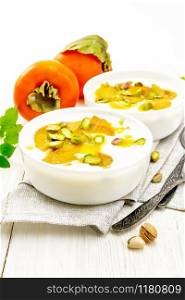 Dessert of yogurt, persimmon and honey with vanilla, cardamom and pistachios in two bowls on a towel, mint and spoon on background of light wooden board