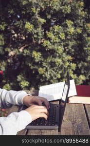 detail of hands typing on laptop with copy space, young latin man working outdoors in park with laptop and books, sitting on wooden bench. detail of hands writing on laptop with copy space