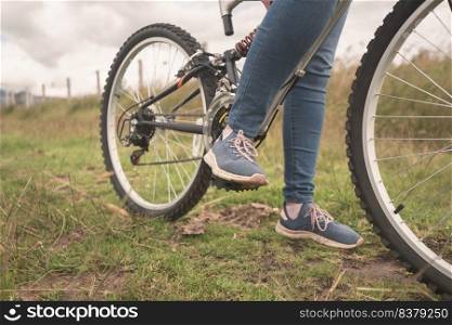Detail of the feet of a young woman wearing blue pants and blue and pink sports shoes on the pedals of her bicycle in the middle of the grassy field on a cloudy day. Detail of the feet of a young woman wearing blue pants on the pedals of her bicycle in the middle of the field on a cloudy day