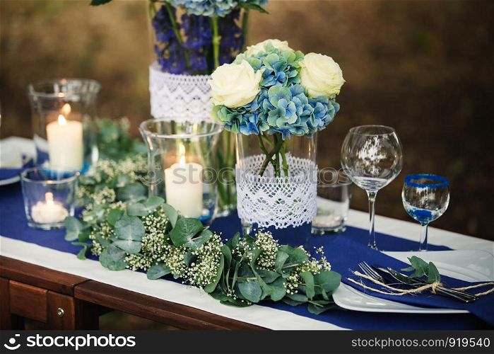 Details. Wedding decorations. On the wooden table in the woods there is a flower arrangement with blue, white, green flowers and greenery, candles, statuettes, glasess, cutlery. photo close-up. Decor. Details. Wedding decorations. On the wooden table in the woods there is a flower arrangement with blue, white, green flowers and greenery, candles, statuettes, glasess, cutlery. photo close-up