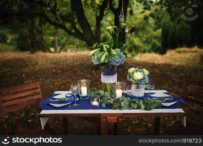 Details. Wedding decorations. On the wooden table in the woods there is a flower arrangement with blue, white, green flowers and greenery, candles, statuettes, glasess, cutlery. photo close-up. Decor. Details. Wedding decorations. On the wooden table in the woods there is a flower arrangement with blue, white, green flowers and greenery, candles, statuettes, glasess, cutlery. photo close-up
