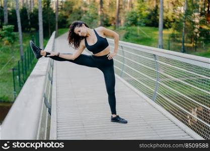 Determined sportswoman stretches legs on bridge, wears active wear, demonstrates good flexibility, poses outdoor against forest background. Woman runner prepares for jogging, has summer workout