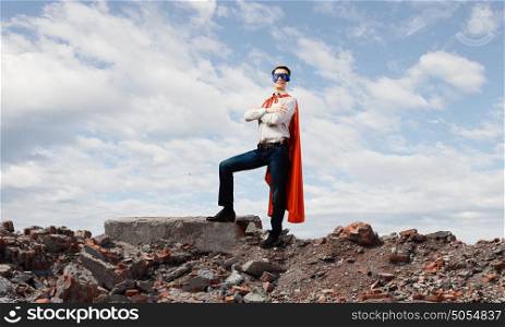 Determined superman. Confident superman in cape and mask standing on ruins