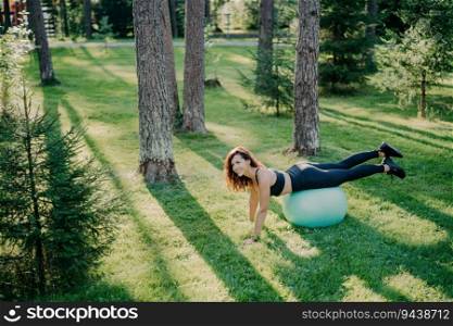 Determined young woman does Pilates in green park, balancing on fitness ball, wearing activewear, happily posing outside. Selective focus.