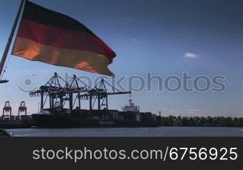 Deutschlandflagge vor blauem Himmel, im Hintergrund KrSne und Containerschiff.