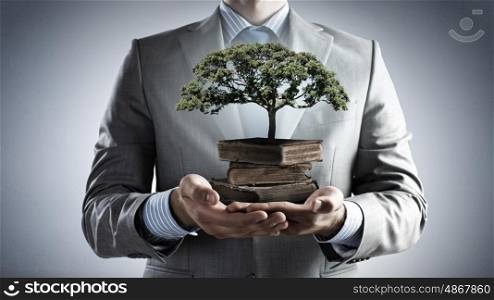 Develope your intelligence. Close up of man holding pile of books with growing green tree in hands