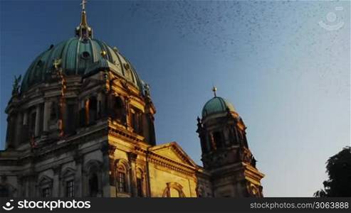 Die Szene zeigt den Berliner Dom gegen Sonnenuntergang. Dabei uberquert ein gewaltiger Schwarm Zugvogel das Gebaude.