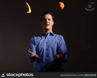 Diet and healthy nutrition. Happy young man juggling with orange and banana tropical fruits on dark gray. Studio shot.