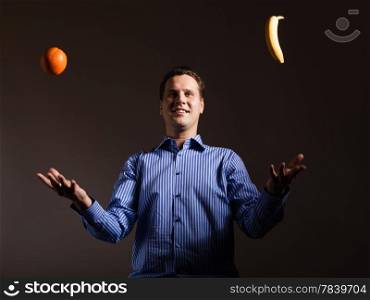 Diet and healthy nutrition. Happy young man juggling with orange and banana tropical fruits on dark gray. Studio shot.