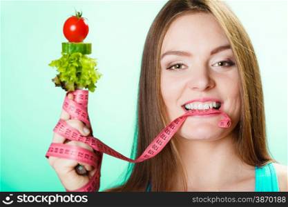 Diet and weight loss concept. Fit woman biting measuring tape with fresh vegetables in hand. Studio shot on blue background.