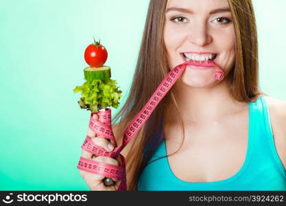 Diet and weight loss concept. Fit woman biting measuring tape with fresh vegetables in hand. Studio shot on green background.