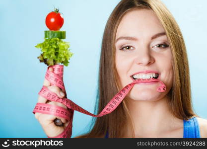 Diet and weight loss concept. Fit woman biting measuring tape with fresh vegetables in hand. Studio shot on blue background.