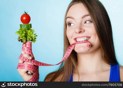 Diet and weight loss concept. Fit woman biting measuring tape with fresh vegetables in hand. Studio shot on blue background.