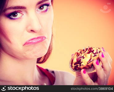 Diet dilemma, grow fat from eating sweets concept. Sad unhappy woman holds cake cupcake in hand orange background