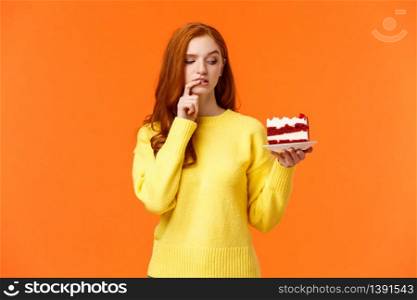 Diet, healthy lifestyle and junk food concept. Cute redhead woman cant resist temptation bite tasty cake, holding dessert on plate and biting lip thinking how many calories in it, orange background.. Diet, healthy lifestyle and junk food concept. Cute redhead woman cant resist temptation bite tasty cake, holding dessert on plate and biting lip thinking how many calories in it, orange background