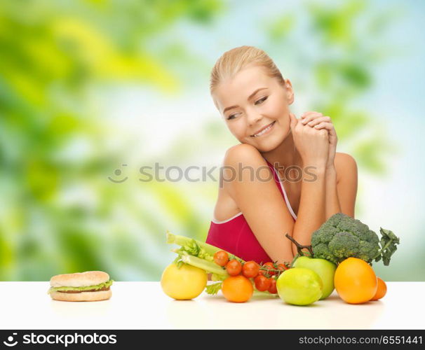 diet, junk food and eating concept - doubting woman with fruits and vegetables looking at hamburger over green natural background. doubting woman with fruits looking at hamburger. doubting woman with fruits looking at hamburger