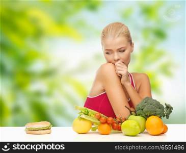 diet, junk food and eating concept - doubting woman with fruits and vegetables looking at hamburger over green natural background. doubting woman with fruits looking at hamburger. doubting woman with fruits looking at hamburger