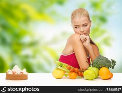 diet, junk food and eating concept - doubting woman with fruits and vegetables looking at cake over green natural background. doubting woman with fruits looking at cake. doubting woman with fruits looking at cake