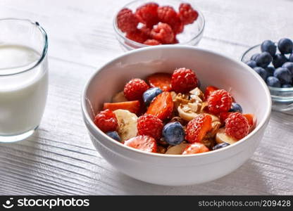 Dietary homemade natural breakfast with fresh organic ingredients - berries, granola, almonds, nuts in a white bowl, soy milk in a glass on a wooden table, copy space. Healthy vegetarian eating.. Natural organic raspberry, strawberry, blueberry, nuts, oat flakes and soy milk - the set of ingredients for healthy breakfast on a wooden background