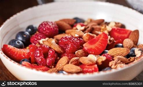 Dietary natural breakfast with fresh organic ingredients - berries, granola, nuts in a white bowl on a wooden table. Healthy vegetarian eating.. Homemade granola in a bowl, strawberries, almonds, blueberries, raspberry, honey - ingredients for natural breakfast.