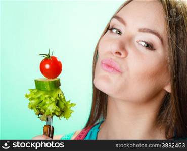 Dieting, healthcare and weight loss concept. Sporty girl fitness woman with measuring tape on neck and holding fork with fresh mixed vegetables on green blue background. Studio shot.