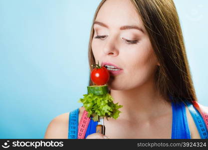 Dieting, healthcare and weight loss concept. Sporty girl fitness woman with measuring tape on neck and holding fork with fresh mixed vegetables on blue background. Studio shot.