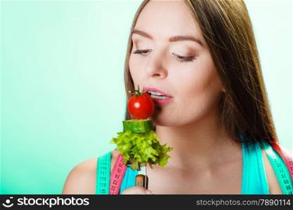 Dieting, healthcare and weight loss concept. Sporty girl fitness woman with measuring tape on neck and holding fork with fresh mixed vegetables on blue background. Studio shot.