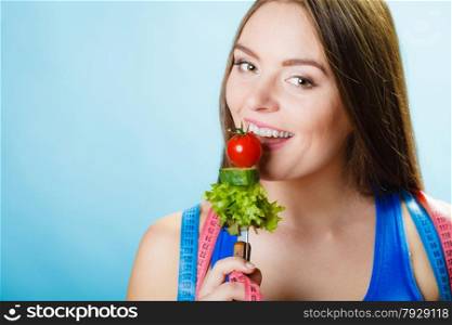 Dieting, healthcare and weight loss concept. Sporty girl fitness woman with measuring tape on neck holding fork with fresh mixed vegetables on blue background. Studio shot.
