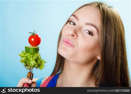 Dieting, healthcare and weight loss concept. Sporty girl fitness woman with measuring tape on neck and holding fork with fresh mixed vegetables on blue background. Studio shot.