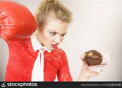Dieting, weight loss concept. Funny blonde woman holding yummy chocolate cupcake cake and boxing glove, fighting off bad food. On grey. Woman fighting off bad food, boxing chocolate cake