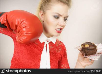 Dieting, weight loss concept. Funny blonde woman holding yummy chocolate cupcake cake and boxing glove, fighting off bad food. On grey. Woman fighting off bad food, boxing chocolate cake