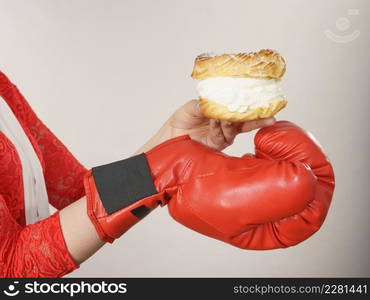 Dieting, weight loss concept. Funny blonde woman holding yummy choux puff cake with whipped cream and boxing glove, fighting off bad food. On grey. Woman fighting off bad food, boxing cream puff cake